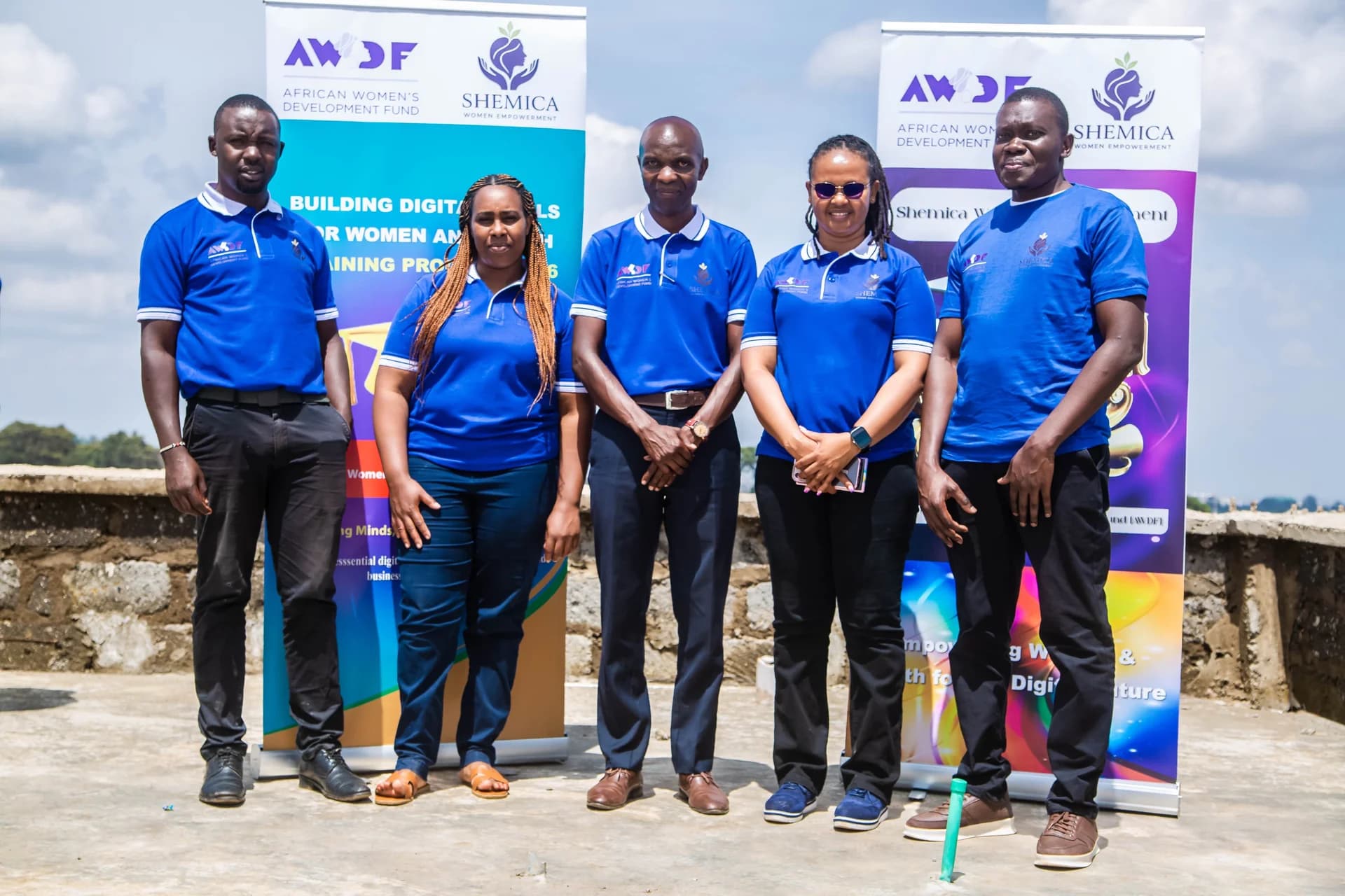 Five people in blue Shemica shirts stand outdoors in front of training banners.