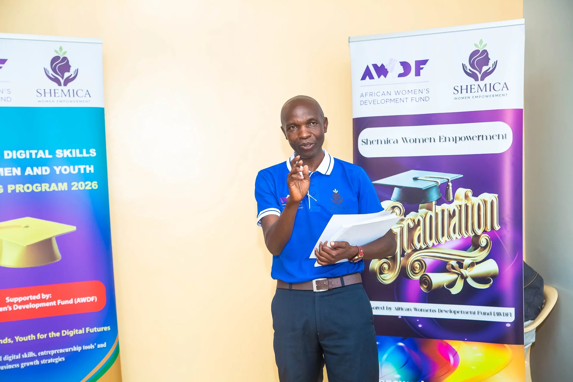 A speaker in a blue shirt addresses a gathering with papers in hand beside graduation banners.