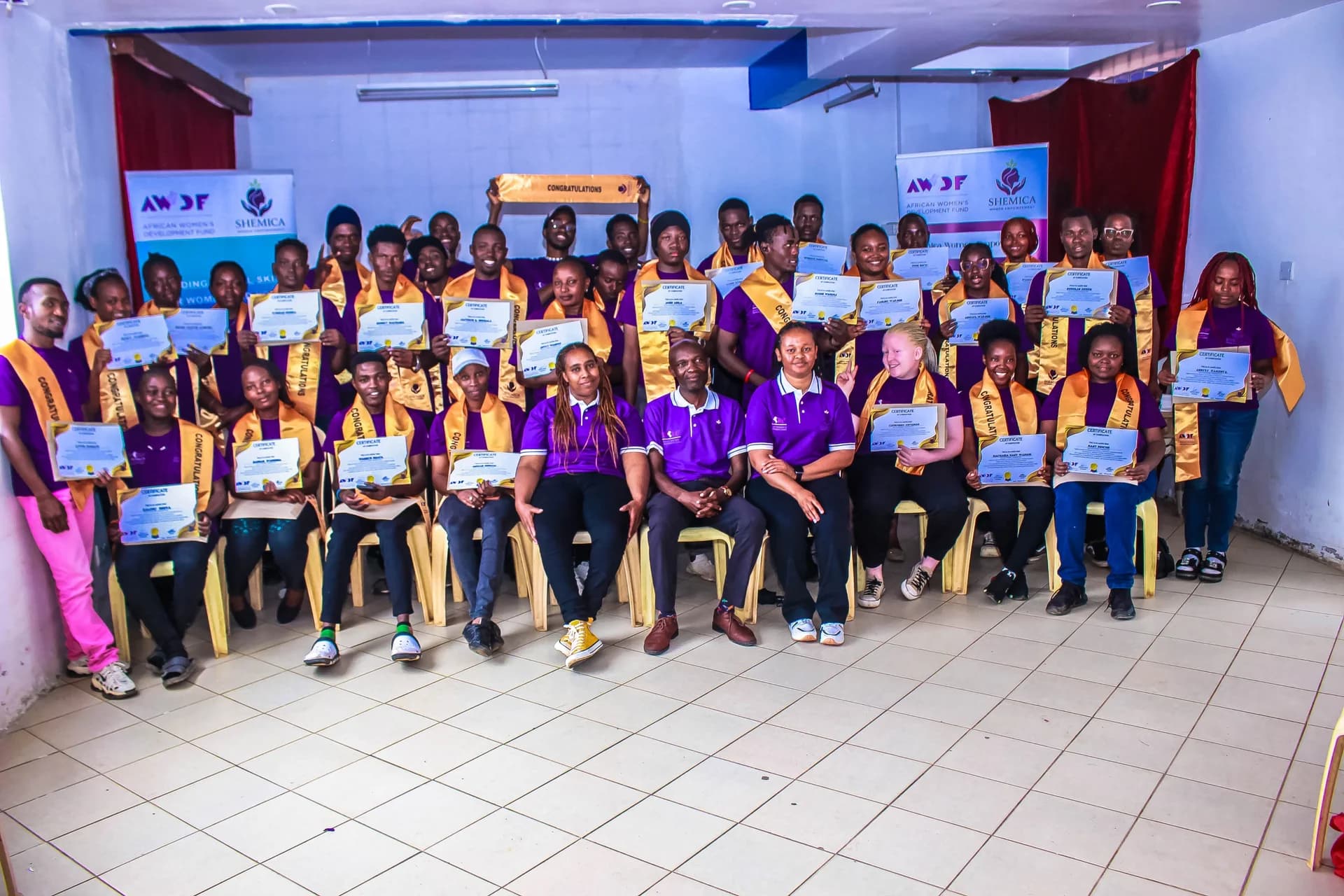 A large group in purple shirts and gold sashes holds certificates in front of Shemica banners.