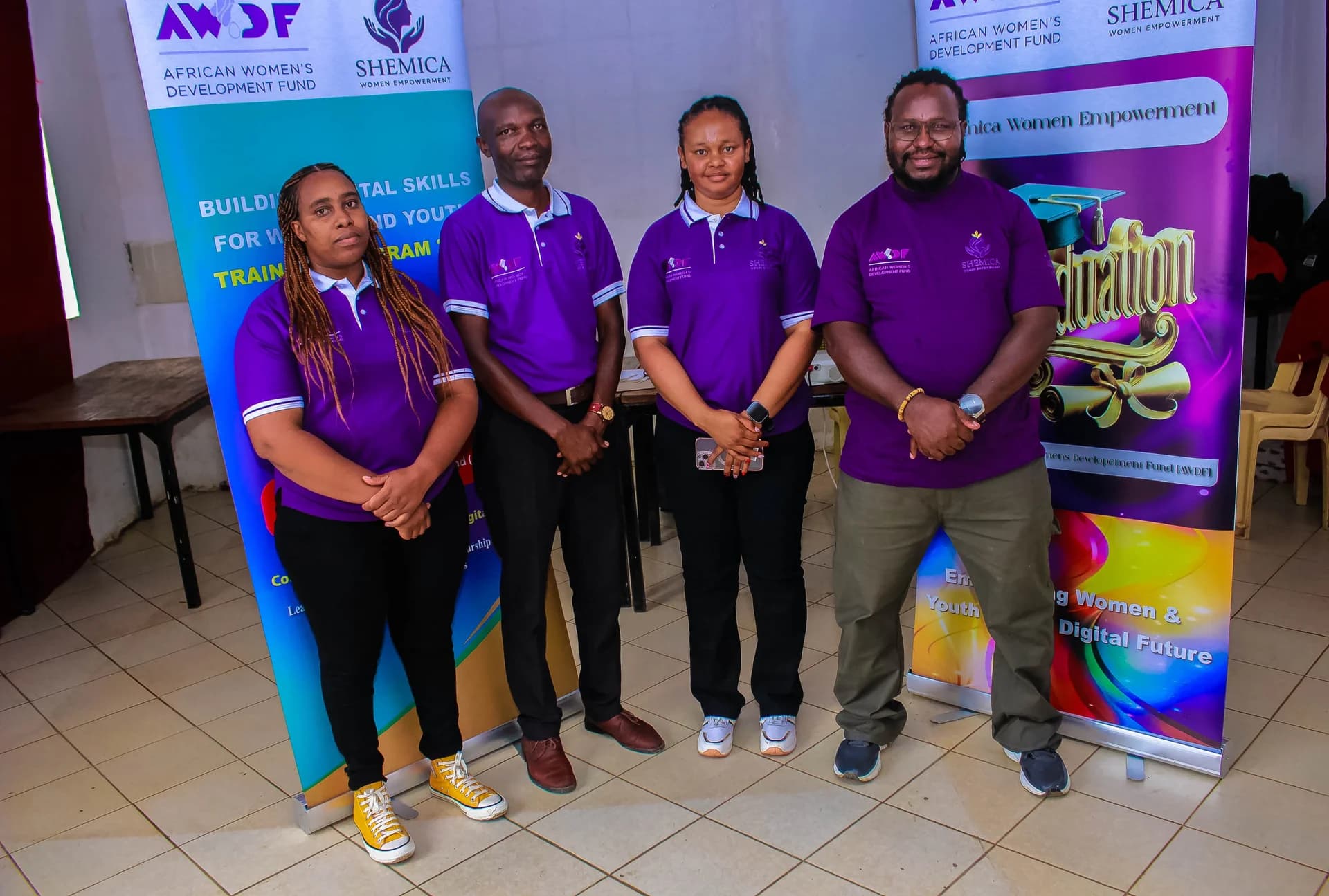 Four people in purple shirts stand together beside Shemica and AWDF banners.