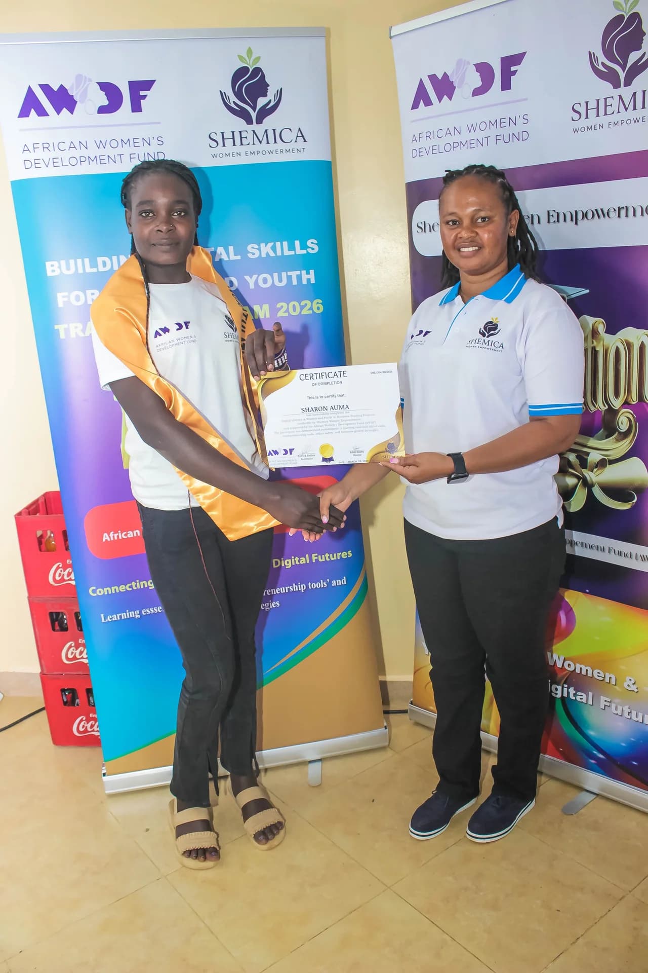 A participant in a white shirt and gold sash holds a certificate alongside a presenter.