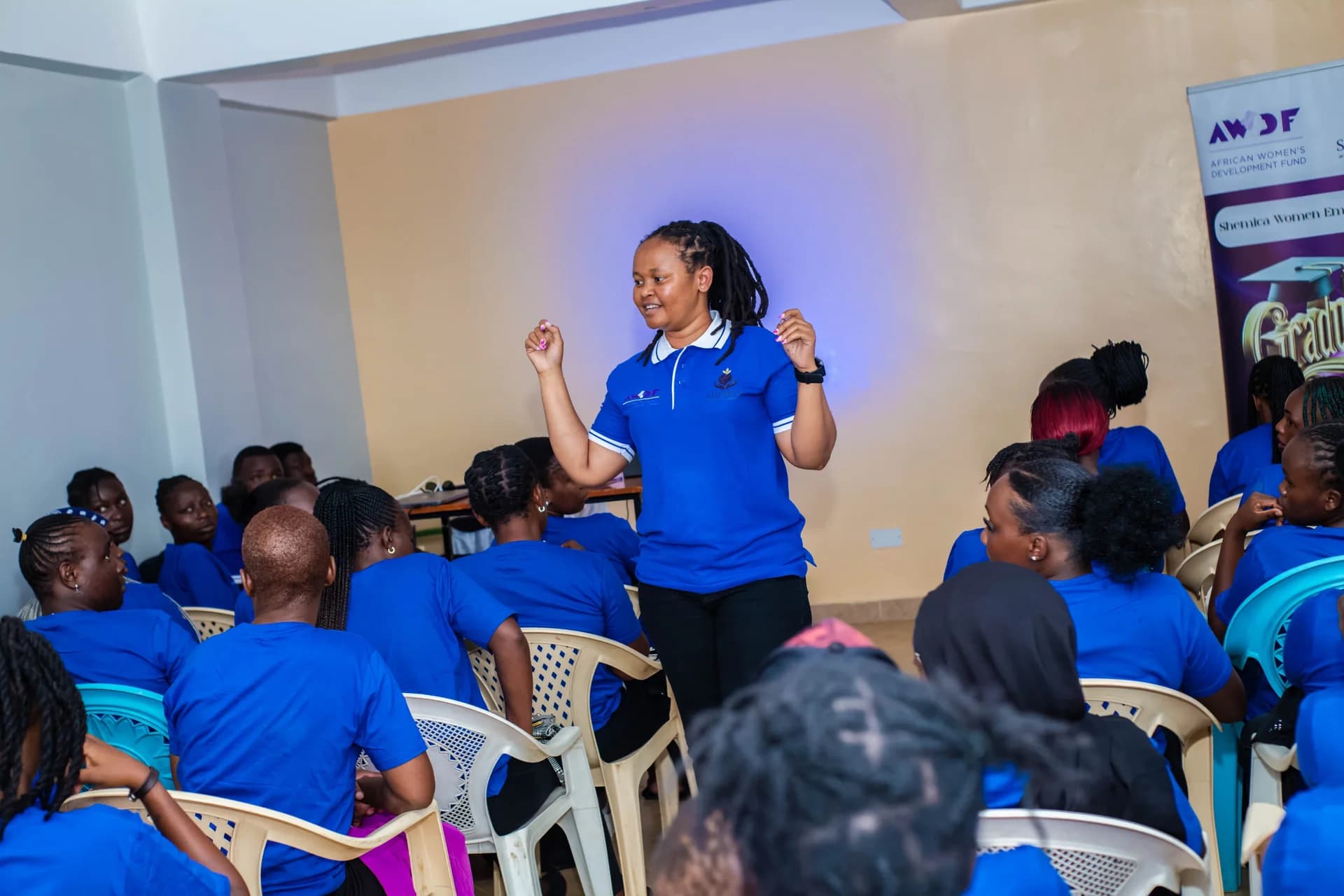 A facilitator in a blue Shemica shirt speaks with a room of seated participants.