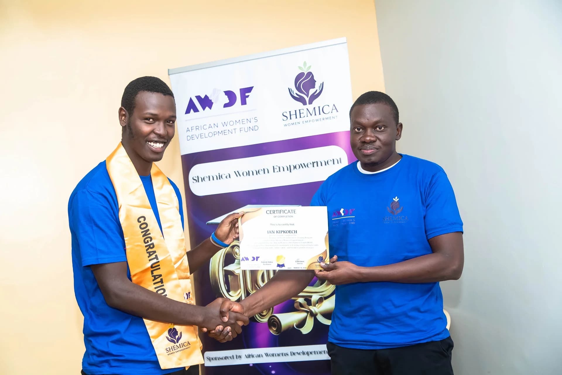 A young man wearing a gold sash holds his certificate and shakes hands with a presenter.