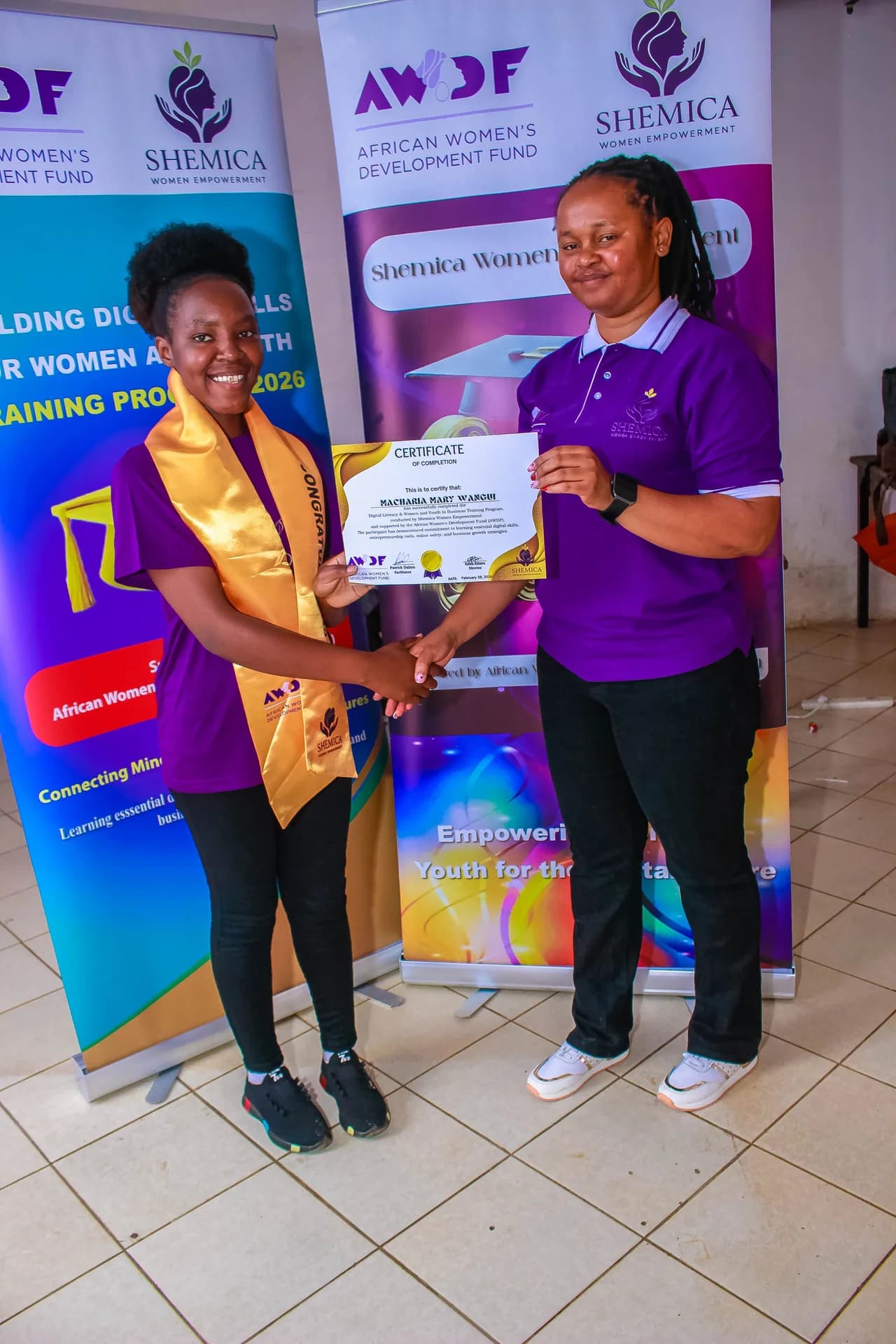 Two women in purple shirts hold a certificate together beside Shemica banners.
